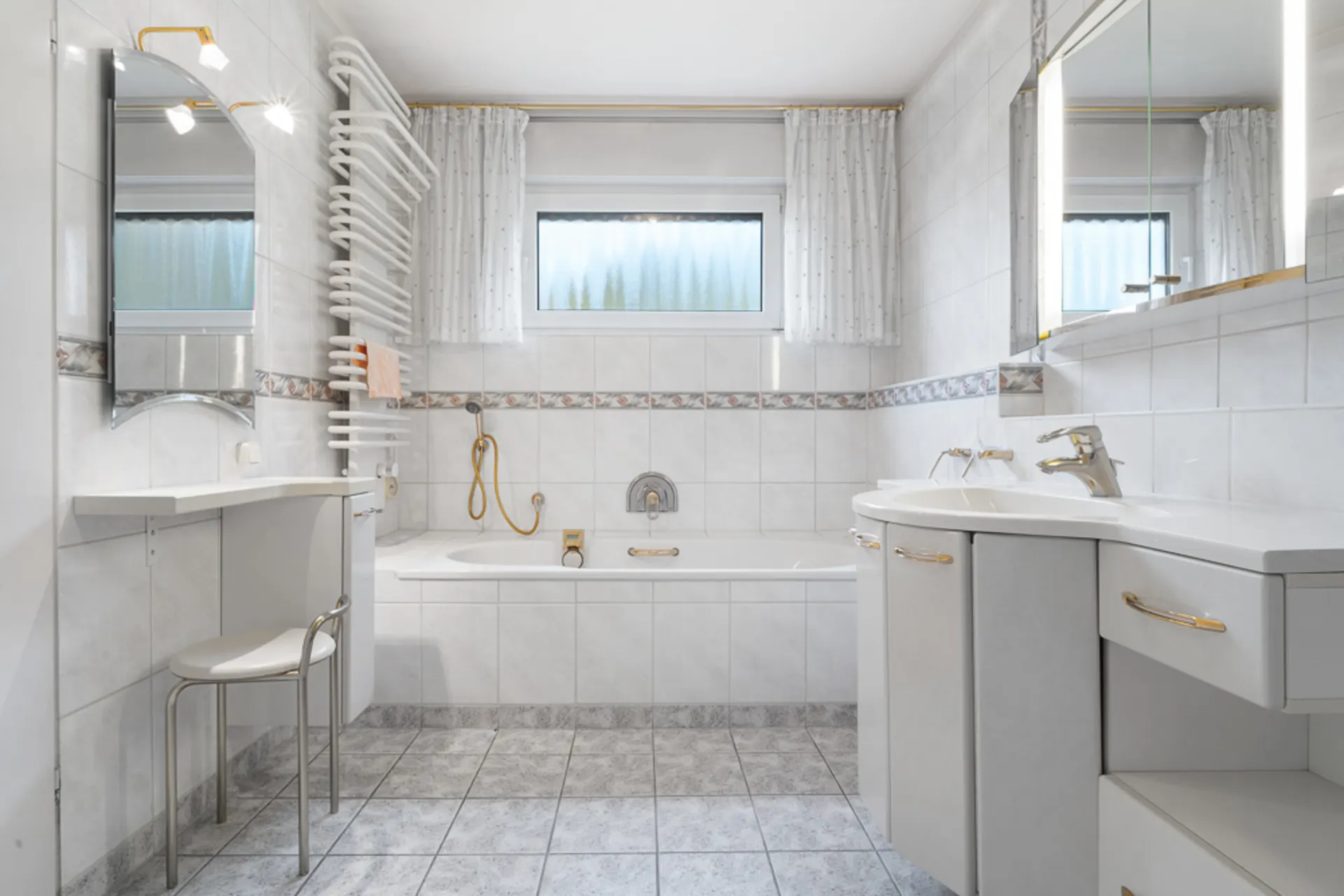 Helles Badezimmer mit Badewanne, zwei Fenstern, Waschbecken und Handtuchheizkörper.
