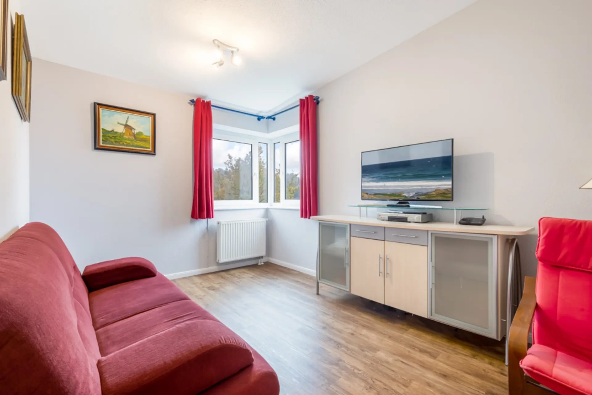 Wohnzimmer mit rotem Sofa, TV auf Sideboard, rotem Sessel und Fenster mit roten Vorhängen.