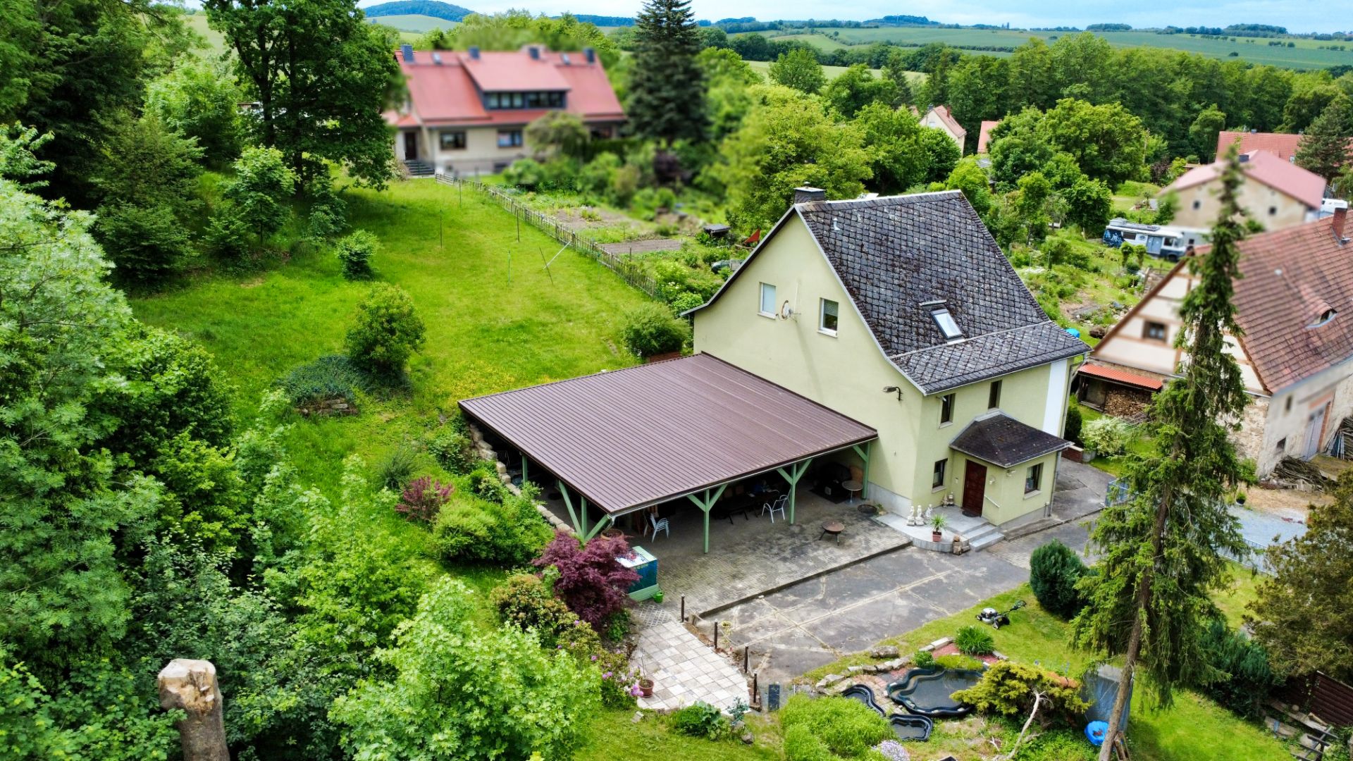 Gelbes Einfamilienhaus mit überdachtem Carport und Garten, umgeben von Bäumen und Wiese.