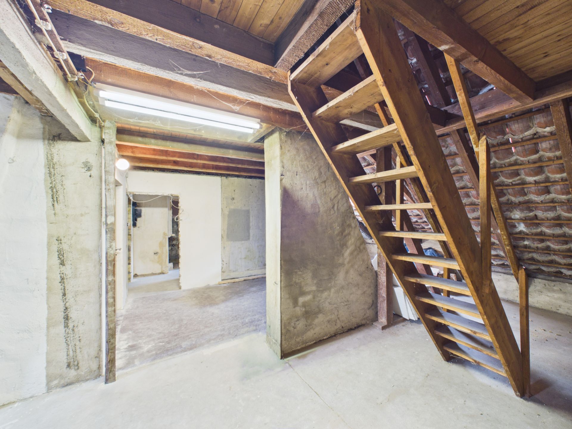 Holztreppe in einem Kellerraum mit Betonwänden und sichtbaren Deckenbalken.