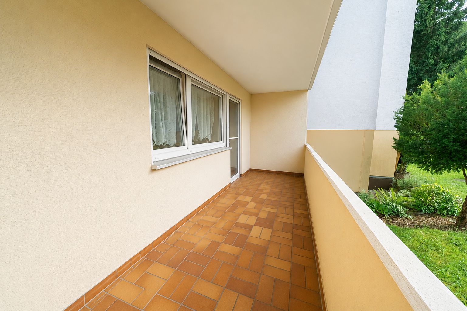 Langer Balkon mit braunen Fliesen, Geländer und Zugang zu einem Gartenbereich.