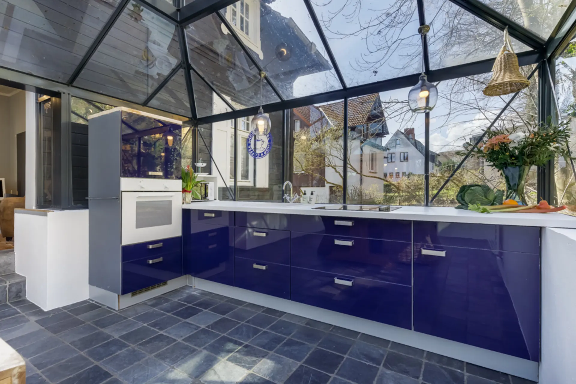 Moderne Küche mit blauen Schränken und Glasdach, Blick auf den Garten und angrenzende Häuser.