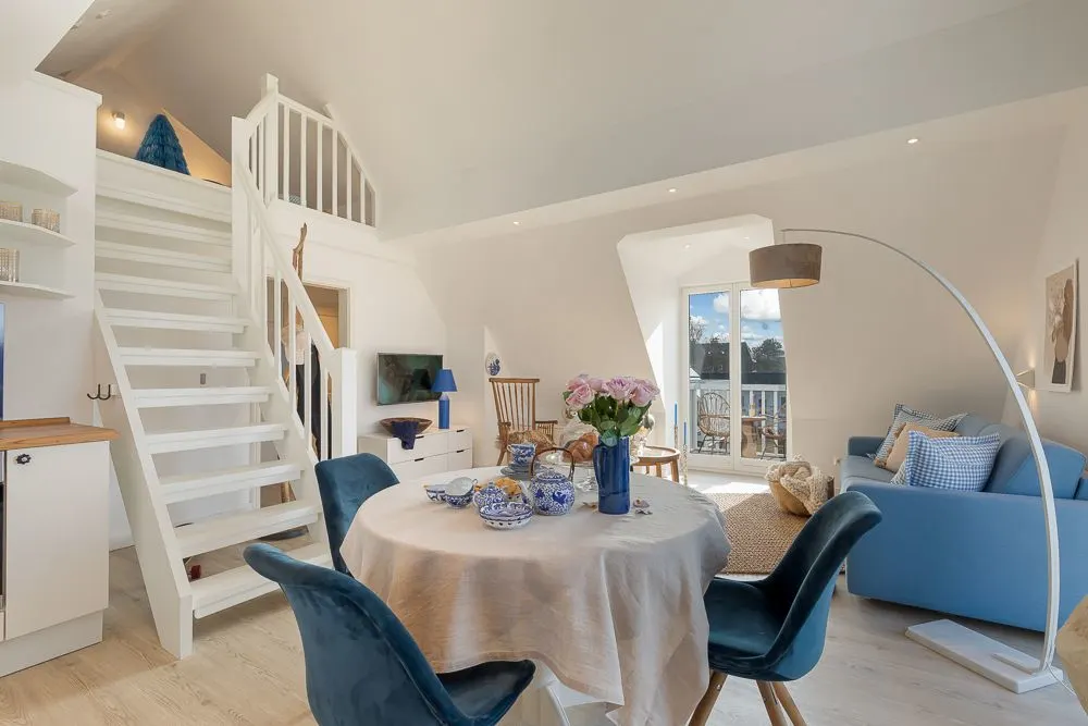 Heller Wohnraum mit Esstisch, blauen Stühlen, Sofa, Treppe und großem Fenster.