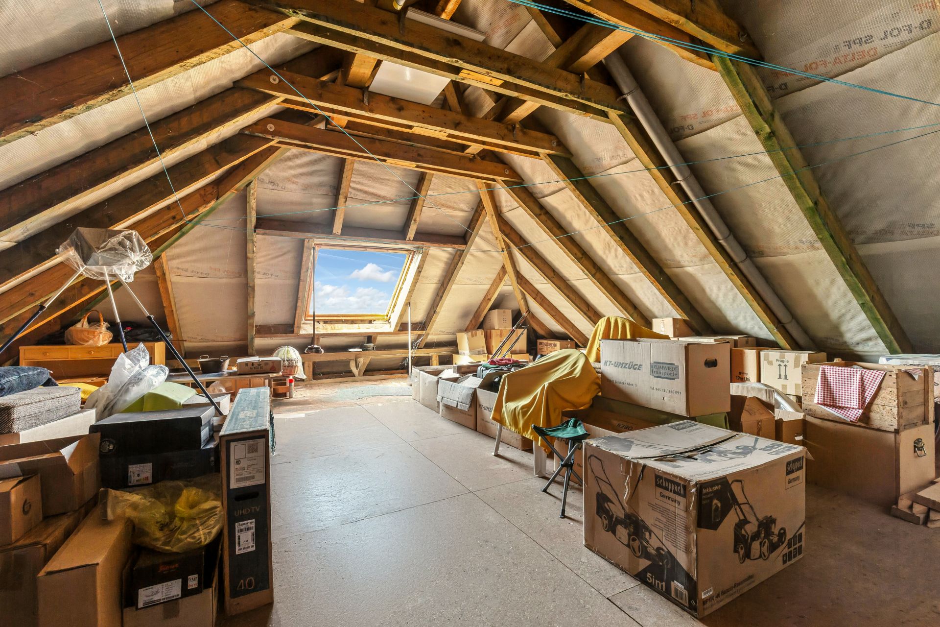Dachboden mit sichtbaren Holzbalken, Dachfenster und mehreren Kartons sowie gelagerten Gegenständen.