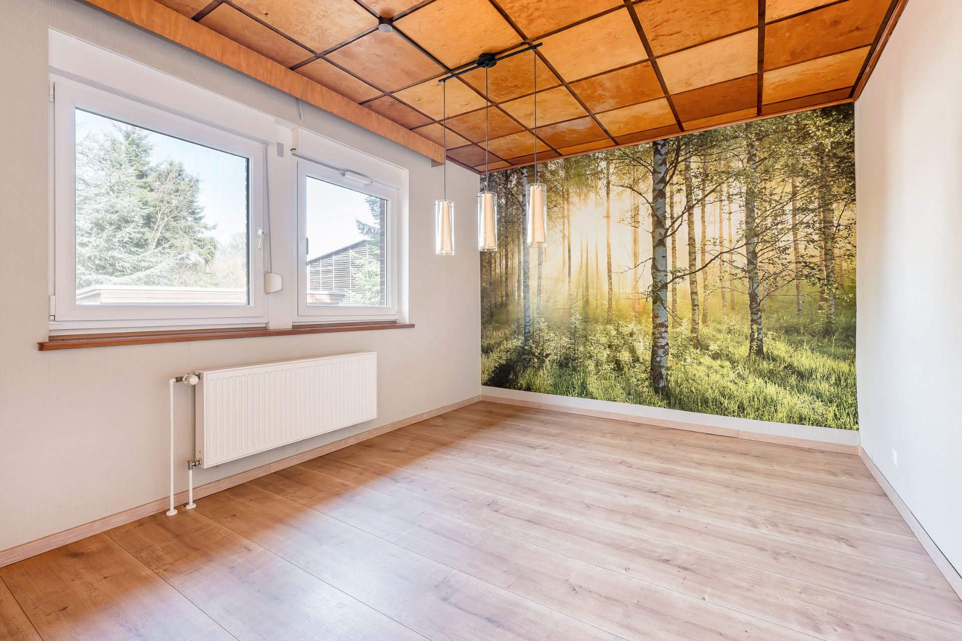 Heller Raum mit Holzdecke, zwei Fenstern und einer Waldfototapete an der Wand.