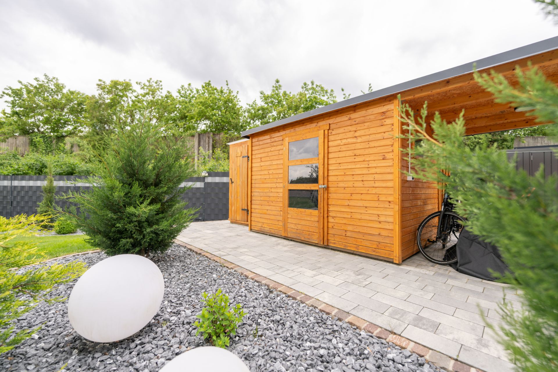 Garten mit Holzhütte, Pflasterweg, Steinen, Büschen und runden Dekokugeln.