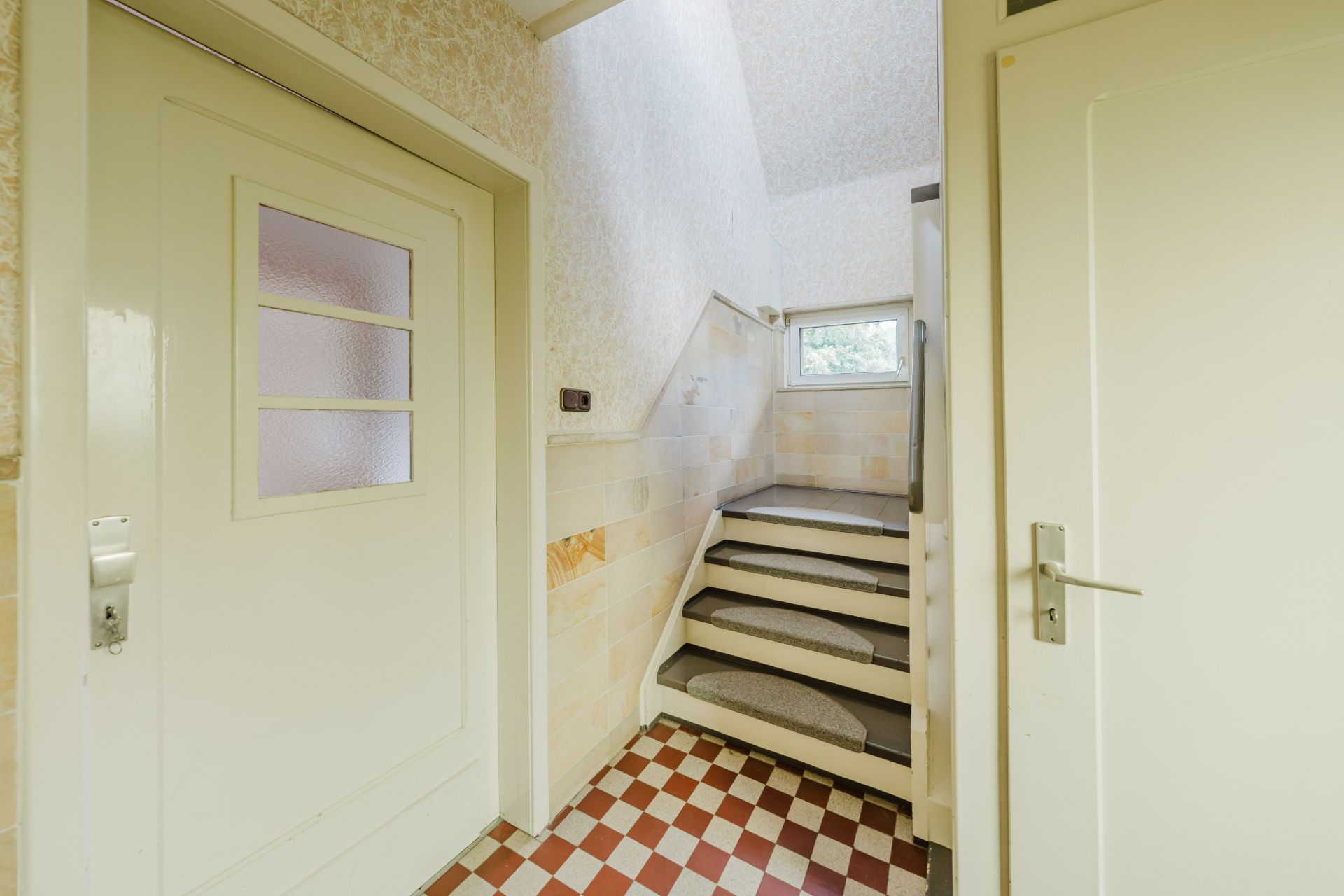 Flur mit gemustertem Fliesenboden, zwei weißen Türen und Treppe mit Fenster.
