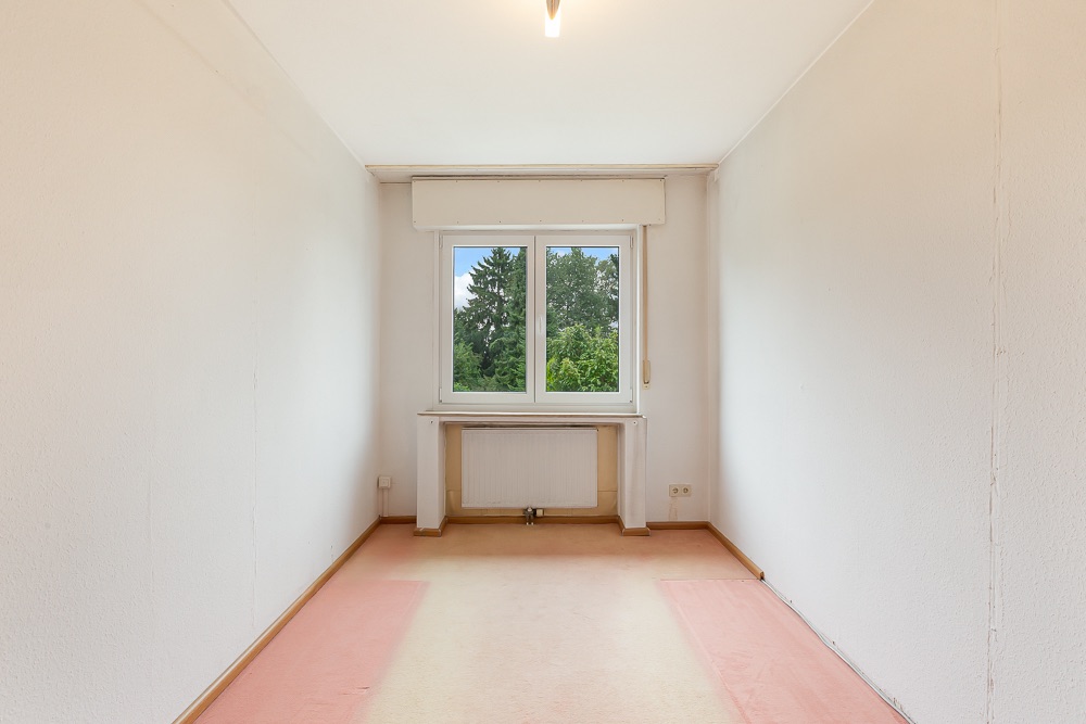 Leeres, schmales Zimmer mit Teppichboden, Fenster und Blick ins Grüne.