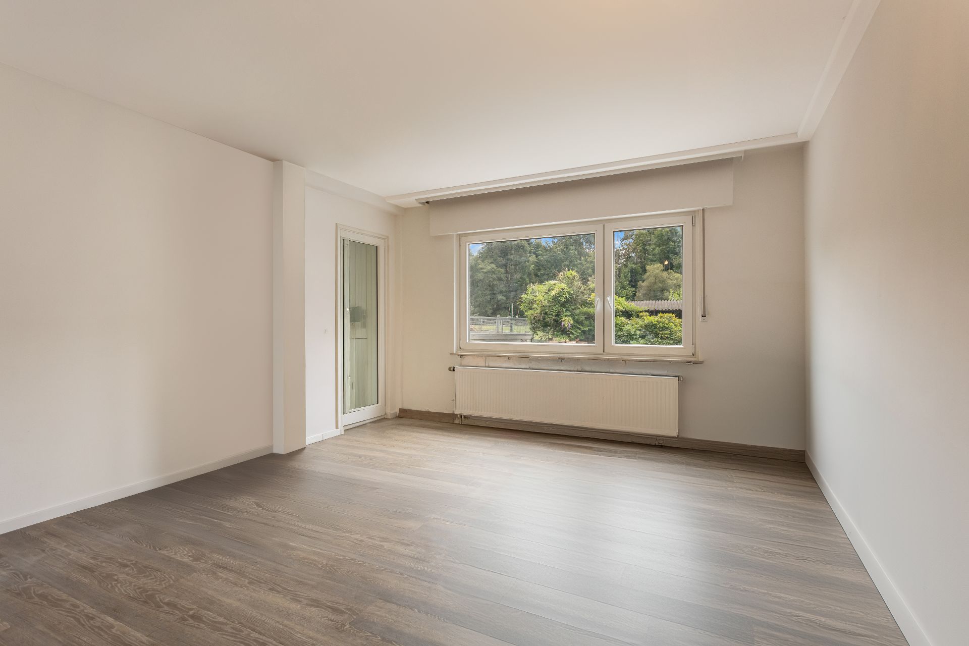 Helles, leeres Zimmer mit Holzboden, großem Fenster und Tür zur Terrasse.