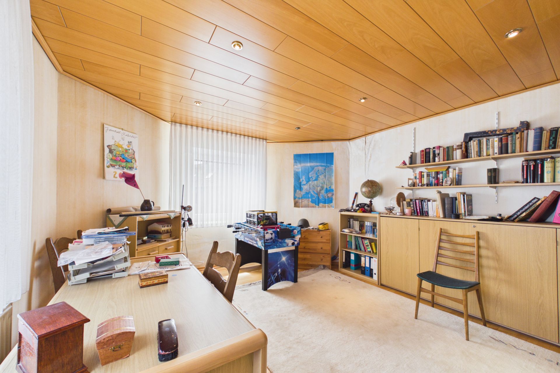 Helles Arbeitszimmer mit Holzdecke, Bücherregalen, Schreibtisch und Stuhl, Fenster mit Vorhängen.
