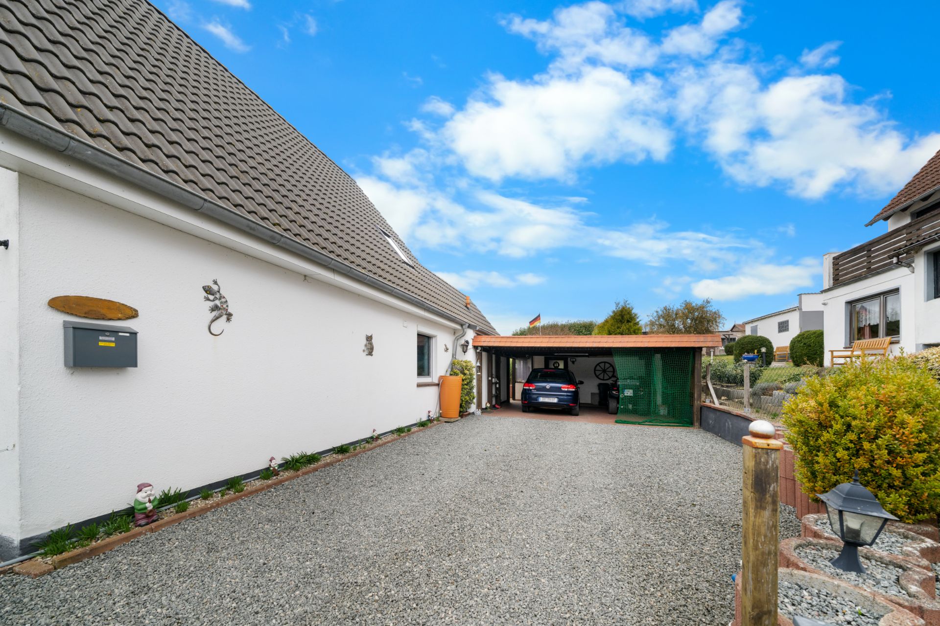 Kiesauffahrt mit Carport, in dem ein Auto steht, neben einem weißen Wohnhaus.
