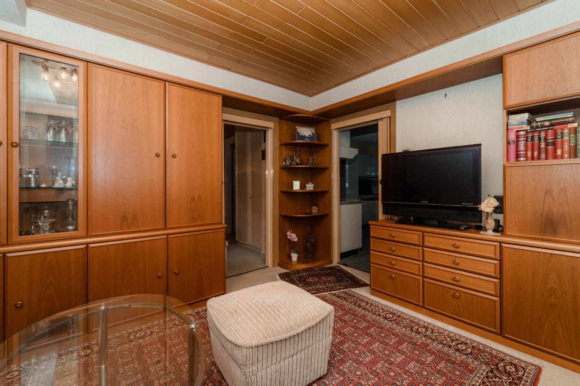 Wohnzimmer mit Holzschränken, kleinem Fernseher, Teppich und Hocker.