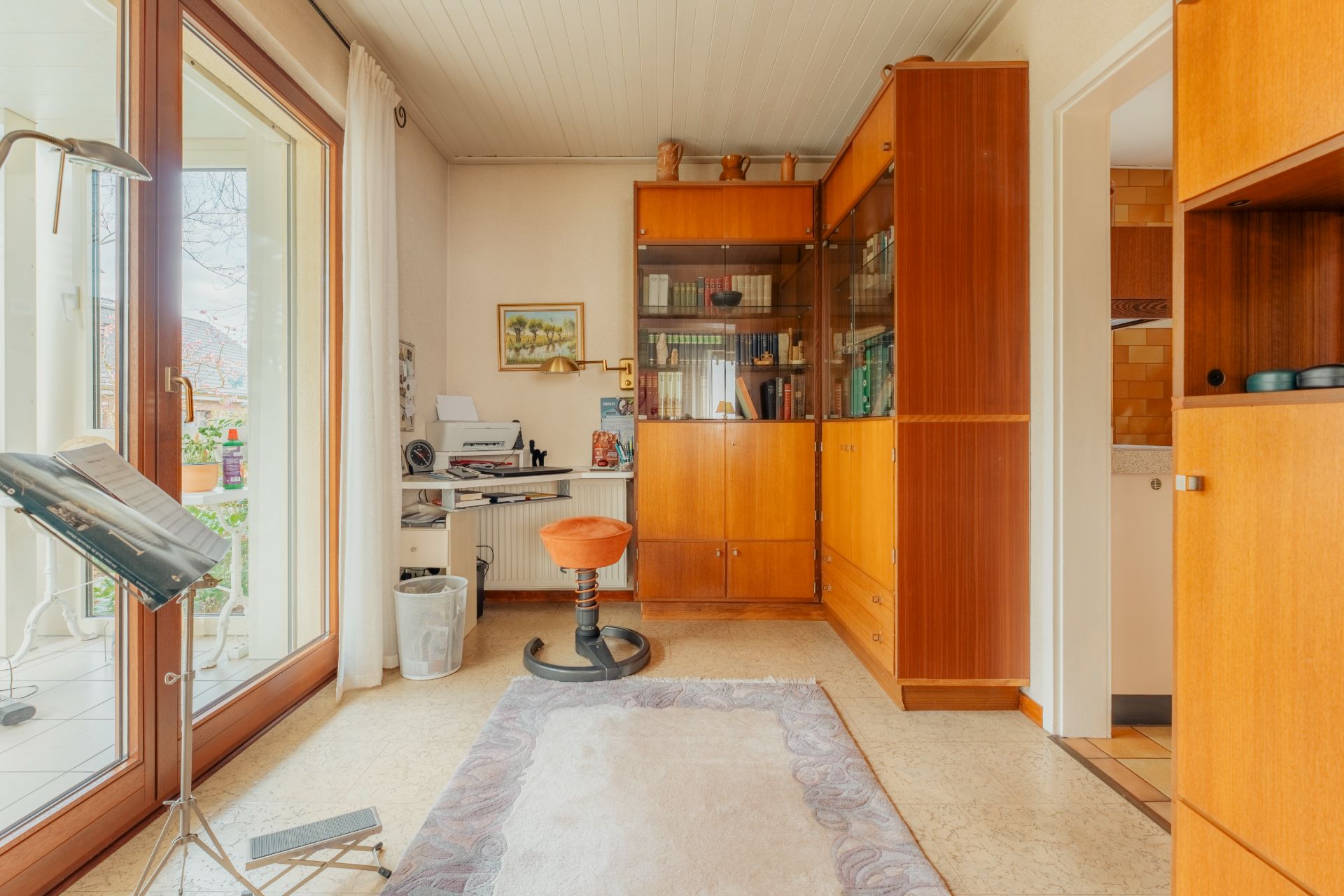 Helles Arbeitszimmer mit großem Fenster, Schreibtisch, Stuhl, Teppich und Holzschränken.