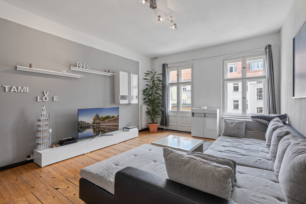 Modernes Wohnzimmer mit grauem Sofa, Fernseher, Couchtisch, großen Fenstern und Holzfußboden.