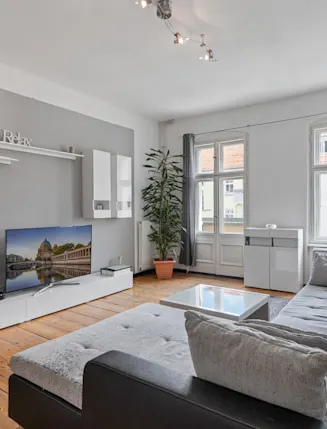 Modernes Wohnzimmer mit grauem Sofa, Fernseher, Couchtisch, großen Fenstern und Holzfußboden.