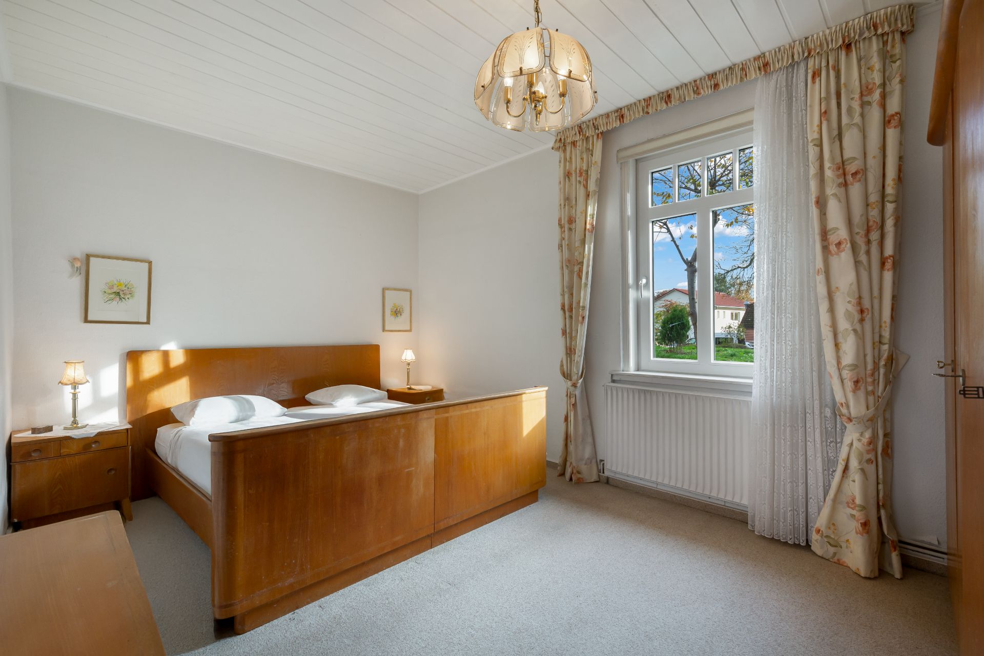 Helles Schlafzimmer mit Holzbett, zwei Nachttischen, Fenster und floralen Vorhängen.