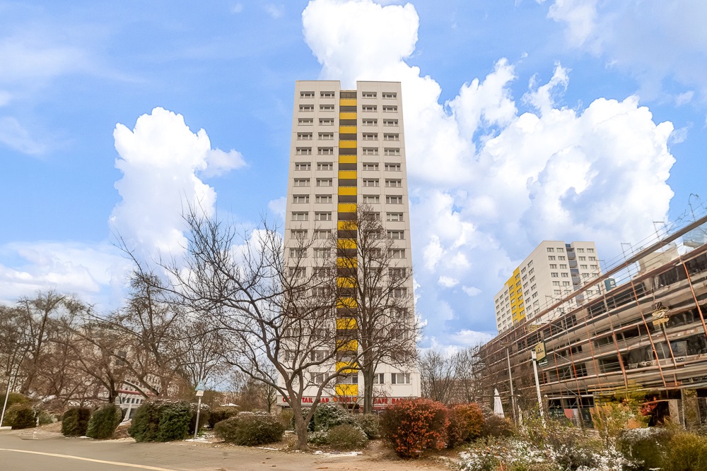 Hohes Wohnhaus mit gelber Fassadenmitte und Bäumen davor, weitere Gebäude im Hintergrund.