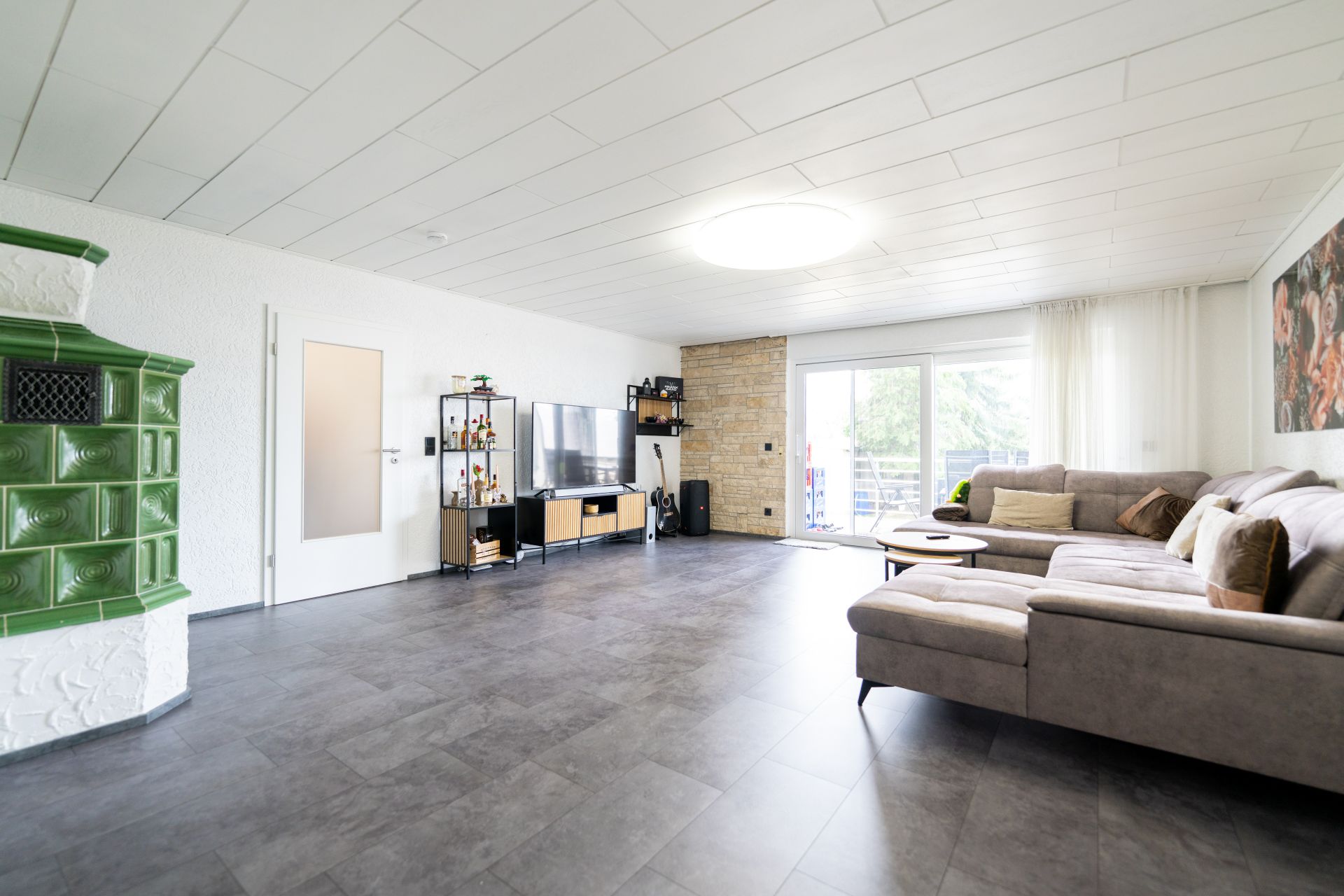 Helles Wohnzimmer mit grauem Ecksofa, grünem Kachelofen, TV und großen Fenstern zu einem Balkon.