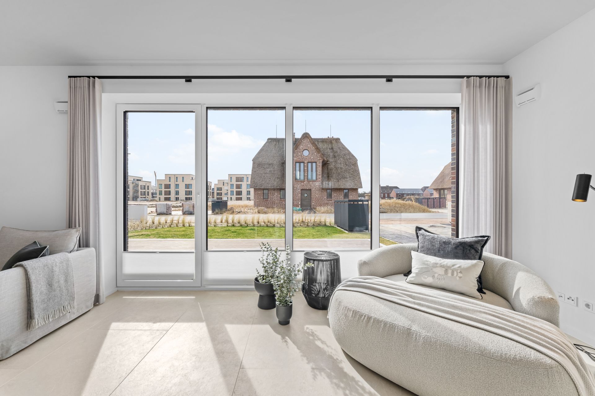 Helles Wohnzimmer mit großem Fenster, Sofa, Sessel und Blick auf Gebäude draußen.