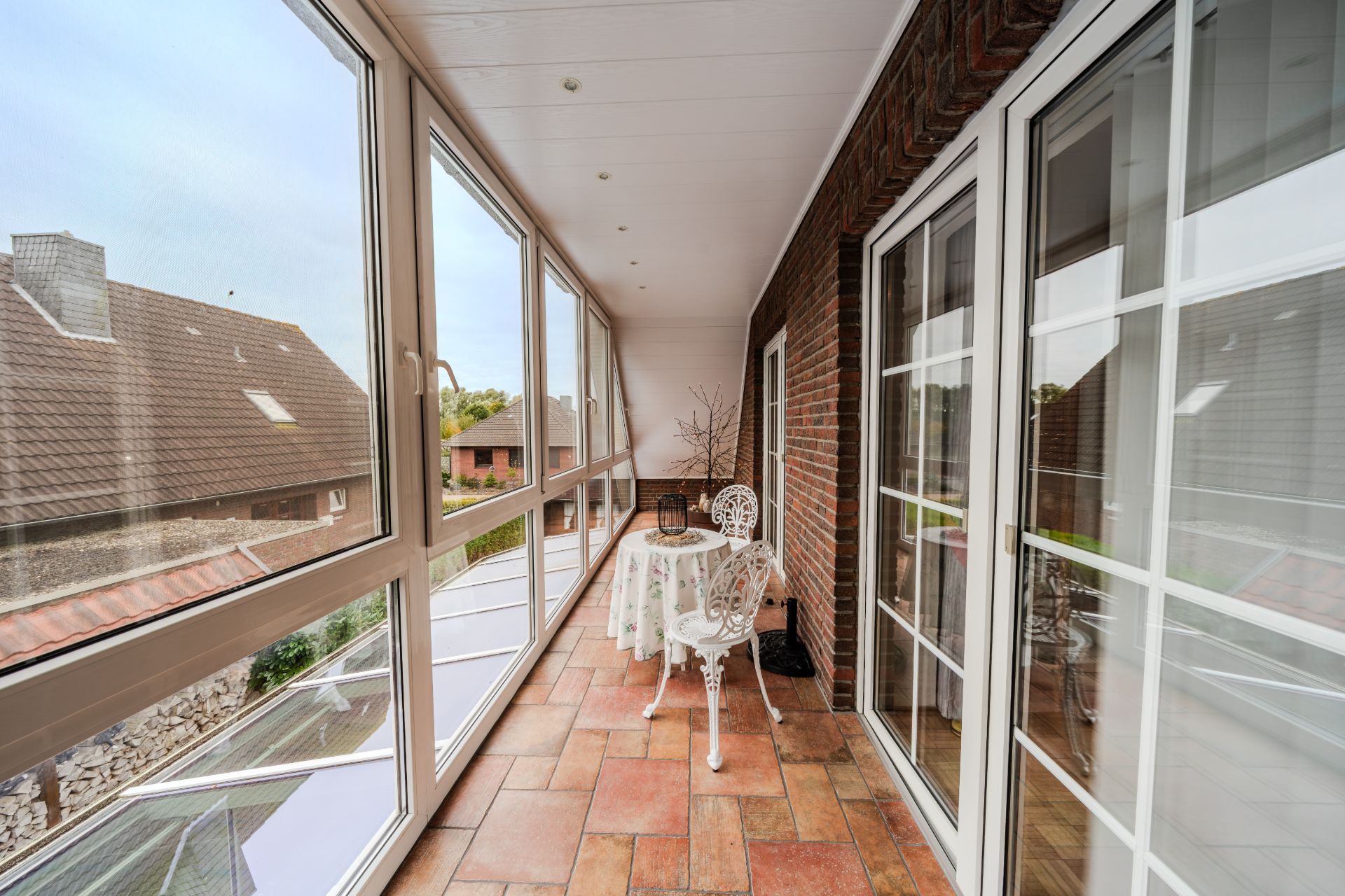 Geschlossene Terrasse mit großen Fenstern, einem kleinen Tisch und zwei weißen Stühlen.