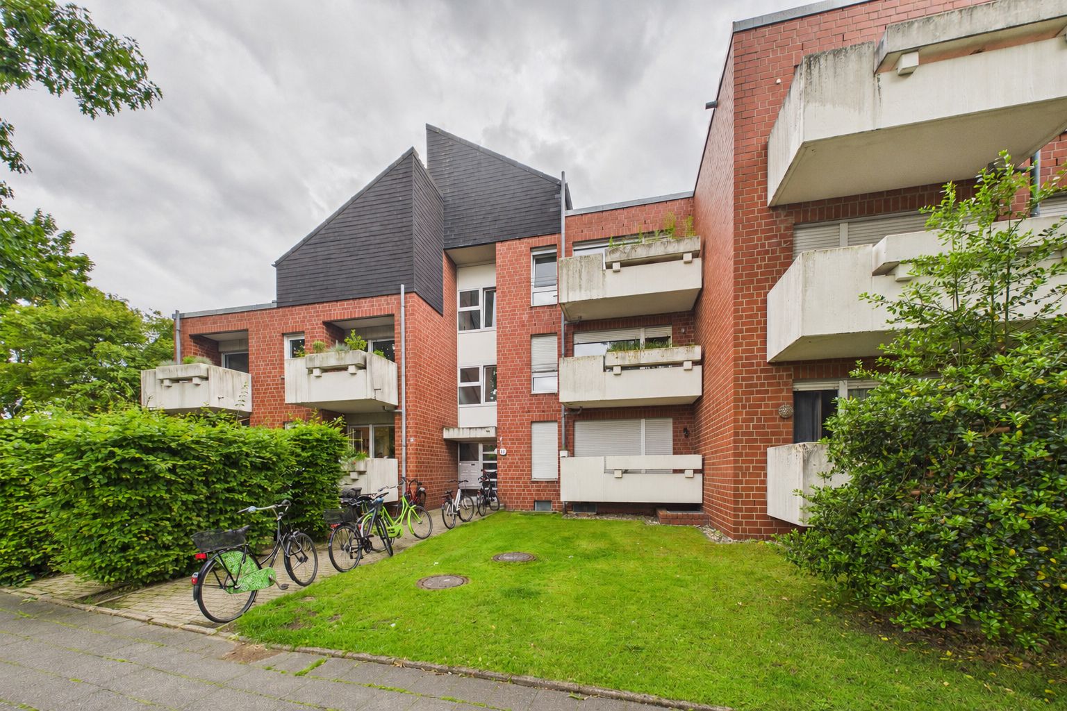 Mehrfamilienhaus mit roten Ziegeln, weißen Balkonen und mehreren Fahrrädern vor dem Eingang.