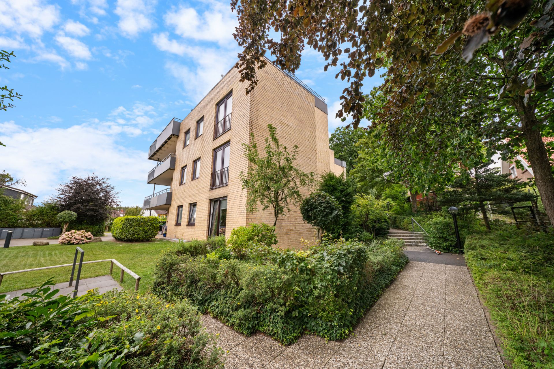 Modernes, dreistöckiges Wohngebäude mit Balkon, umgeben von Garten und grünen Bäumen.