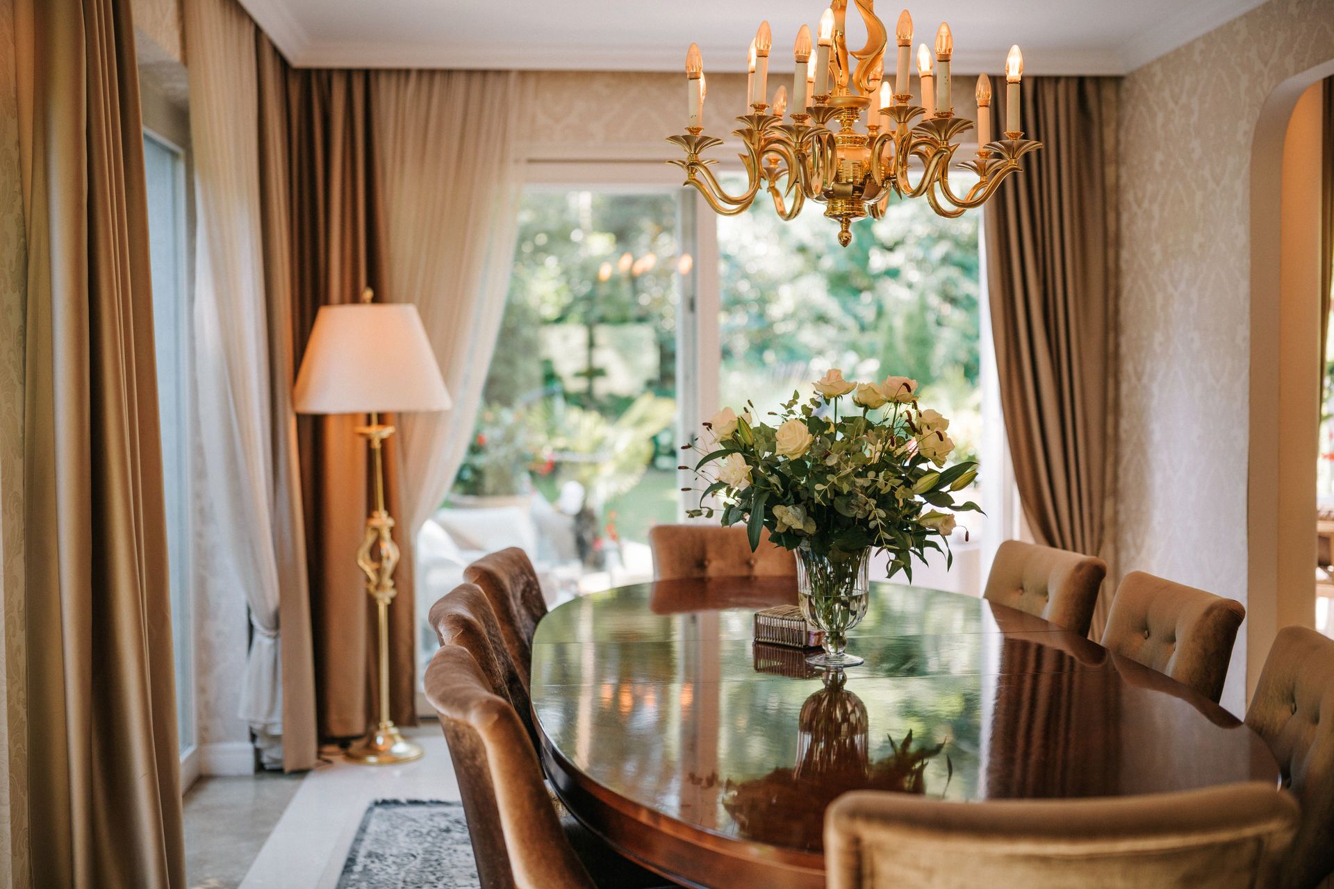 Esszimmer mit ovalem Holztisch, Blumenstrauß, Kronleuchter und großen Fenstern mit Vorhängen.