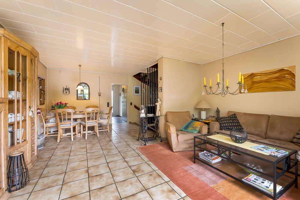 Helles Wohnzimmer mit braunem Sofa, Esstisch, Schrank und Kronleuchter auf Fliesenboden.