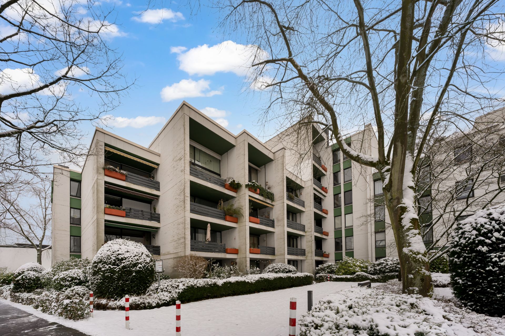 Mehrfamilienhaus mit Balkonen, umgeben von Bäumen und verschneiten Büschen im Winter.