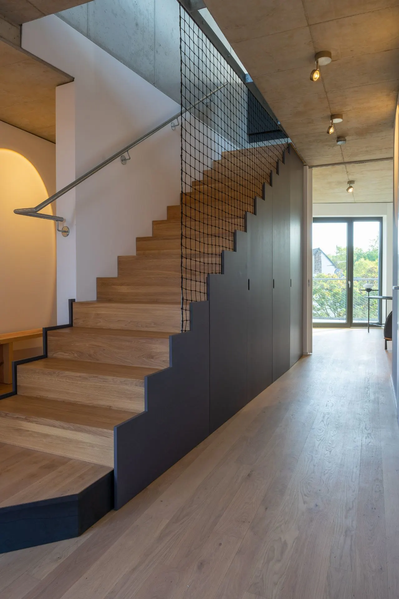 Moderne Holztreppe mit schwarzem Geländer und Netz, heller Flur mit großen Fenstern.