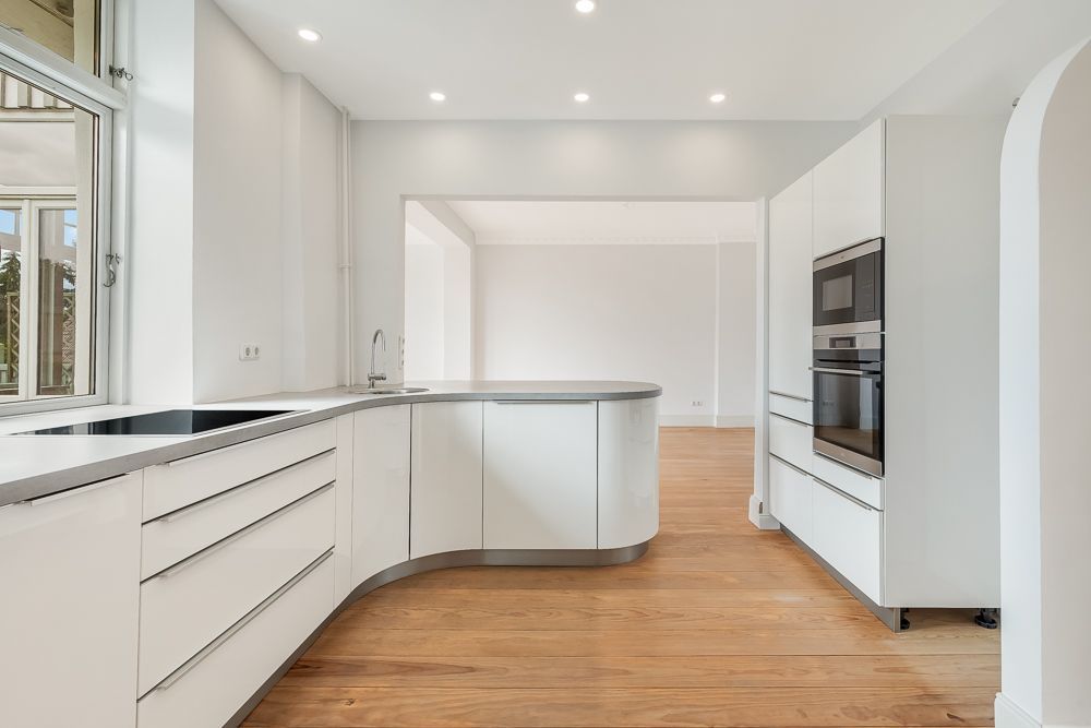 Moderne weiße Einbauküche mit Holzfußboden und großen Fenstern, offene Raumgestaltung.