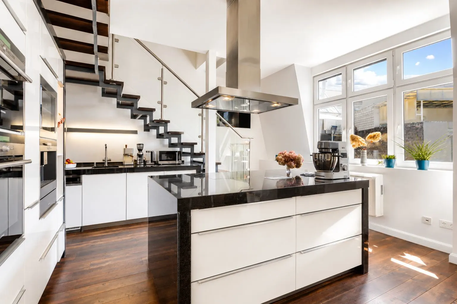 Moderne Küche mit Kochinsel, dunklem Holzboden und Treppe, große Fenster lassen viel Licht herein.