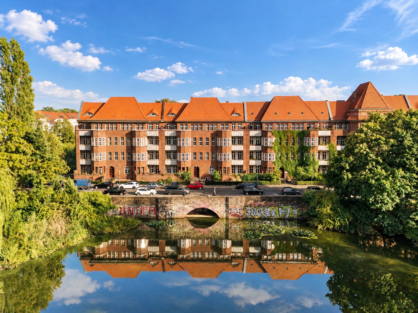 Mehrstöckiges Wohnhaus mit rotem Dach, davor eine Brücke über einen Fluss, umgeben von Bäumen.