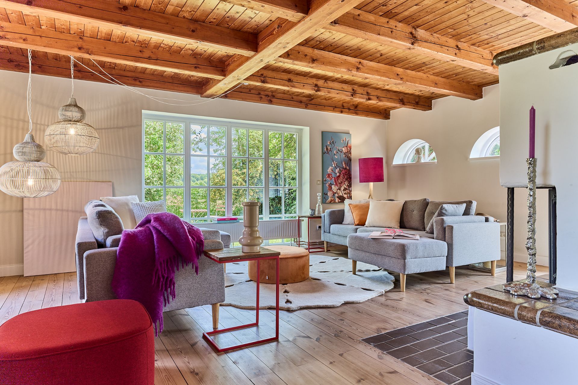 Helles Wohnzimmer mit Holzbalkendecke, großen Fenstern, grauen Sofas und dekorativen Lampen.