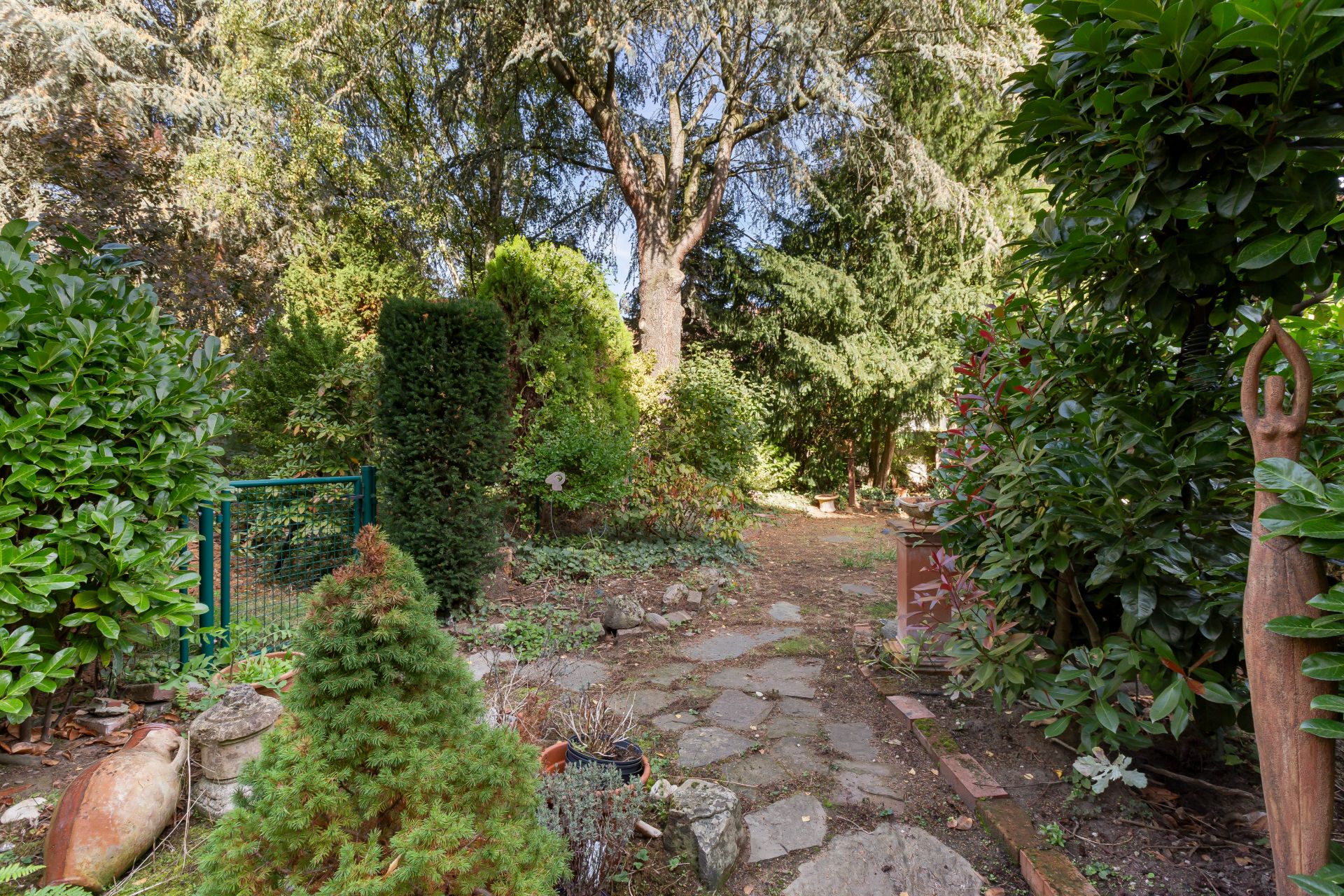 Garten mit Steinweg, Büschen, Bäumen und einer kleinen Statue am rechten Bildrand.