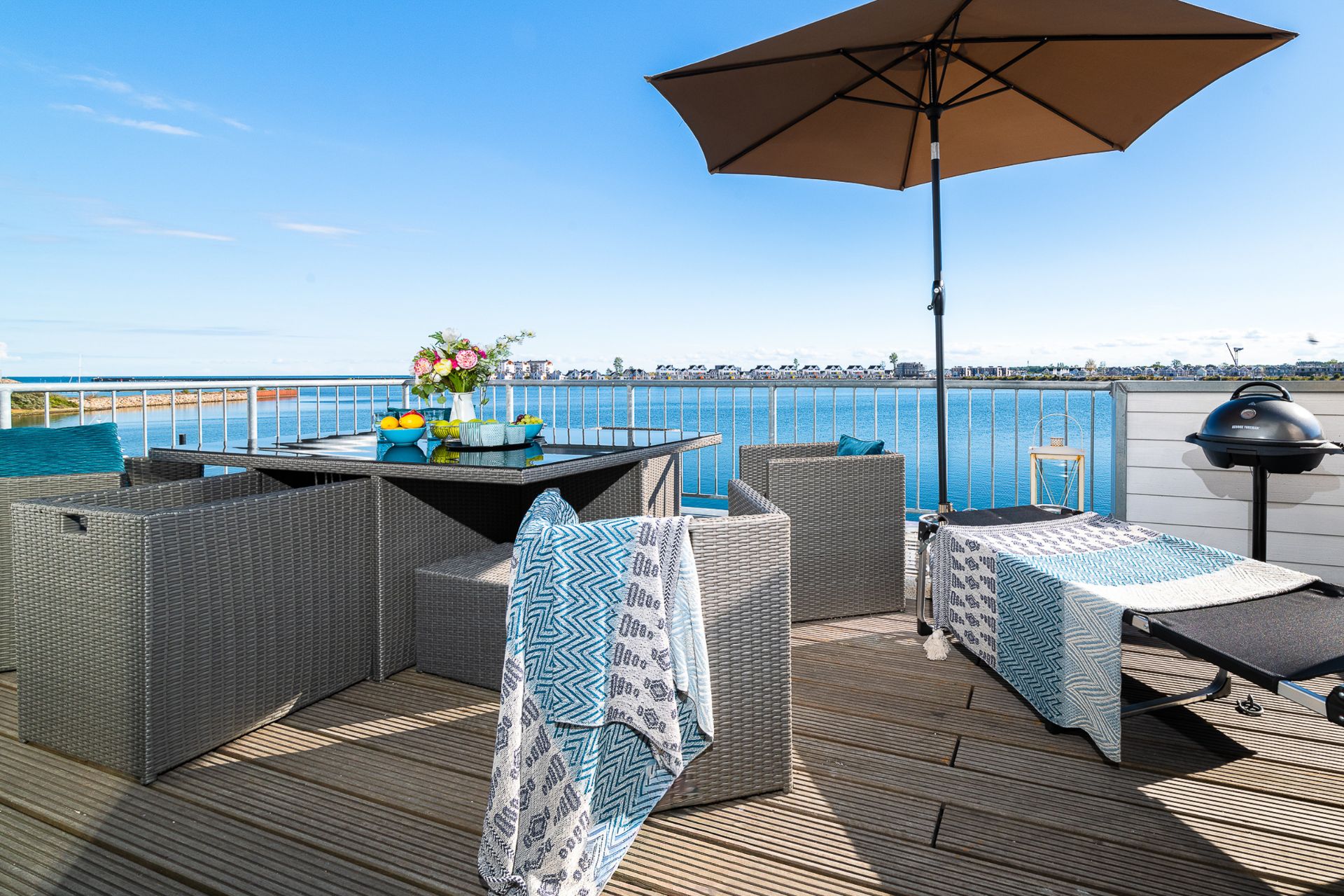 Terrasse mit Sitzgruppe, Liege, Sonnenschirm und Blick auf einen See.