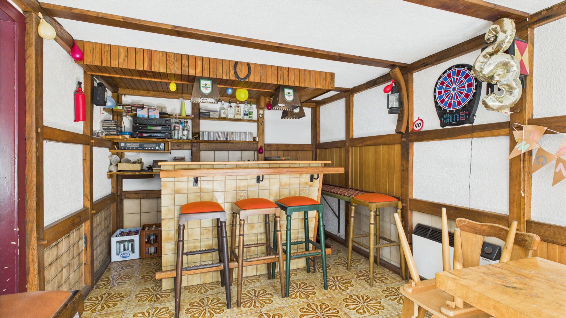 Kleine Hausbar mit Tresen, Hockern, Wandregalen und einer Dartscheibe an der Wand.