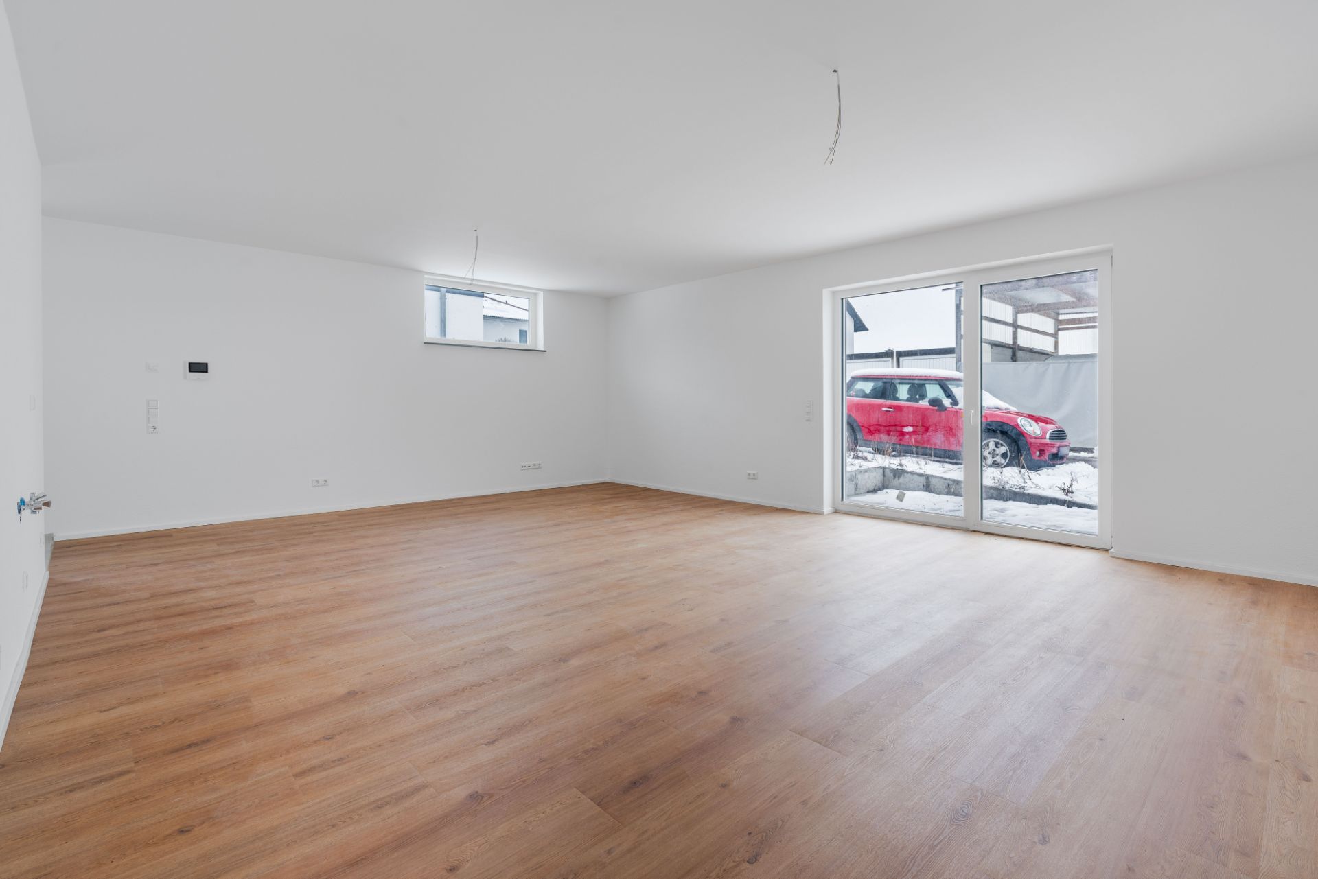 Leerer Raum mit Holzboden, weißer Wand und großen Fenstern zu einer Terrasse mit rotem Auto.