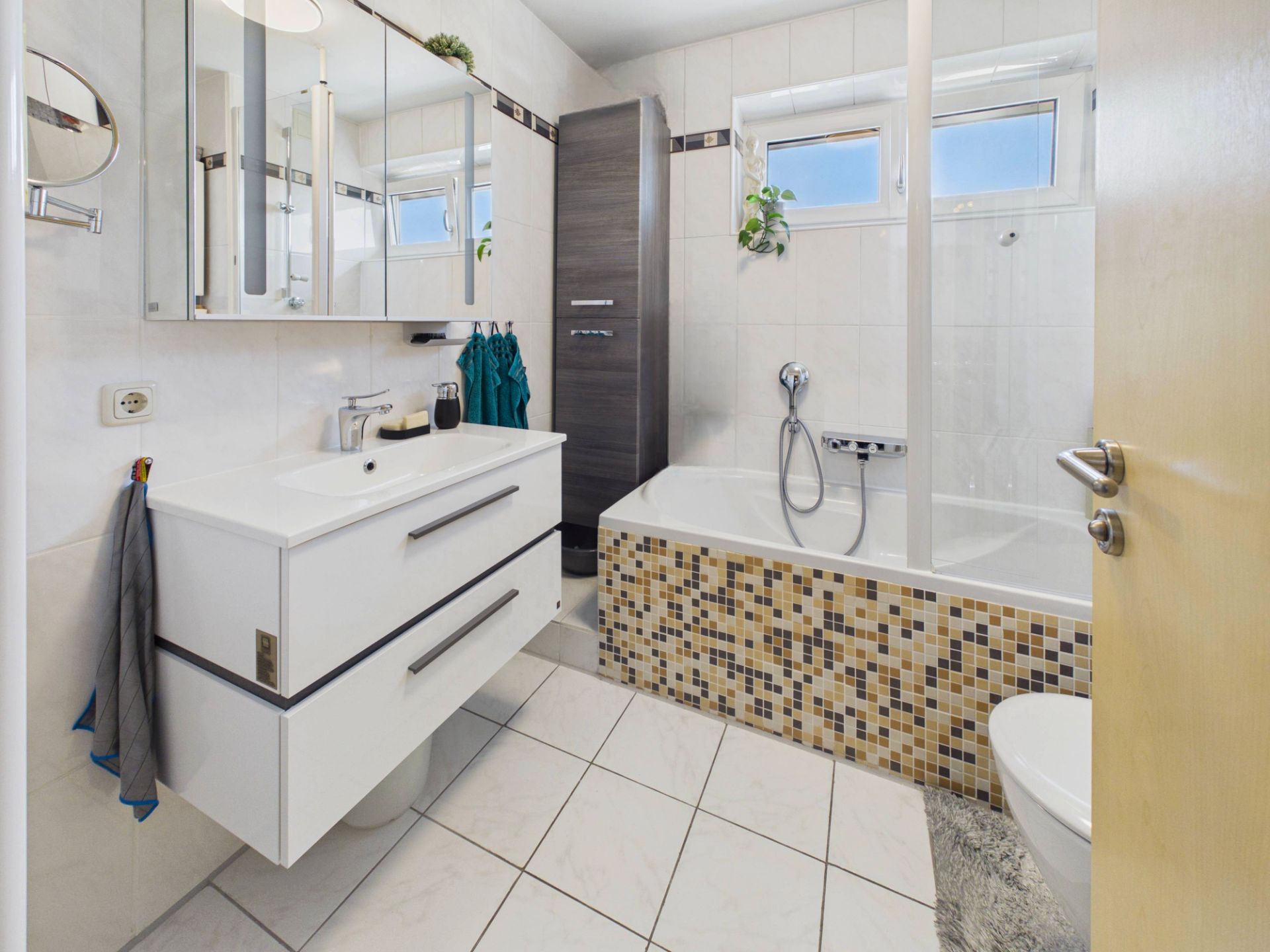 Modernes Badezimmer mit Badewanne, Waschbecken, Spiegelschrank und zwei Fenstern.