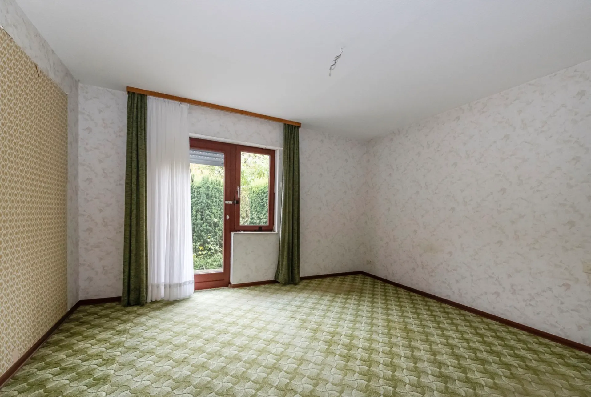 Leeres Zimmer mit gemustertem Teppich, Fenster und Tür mit grünen Vorhängen.