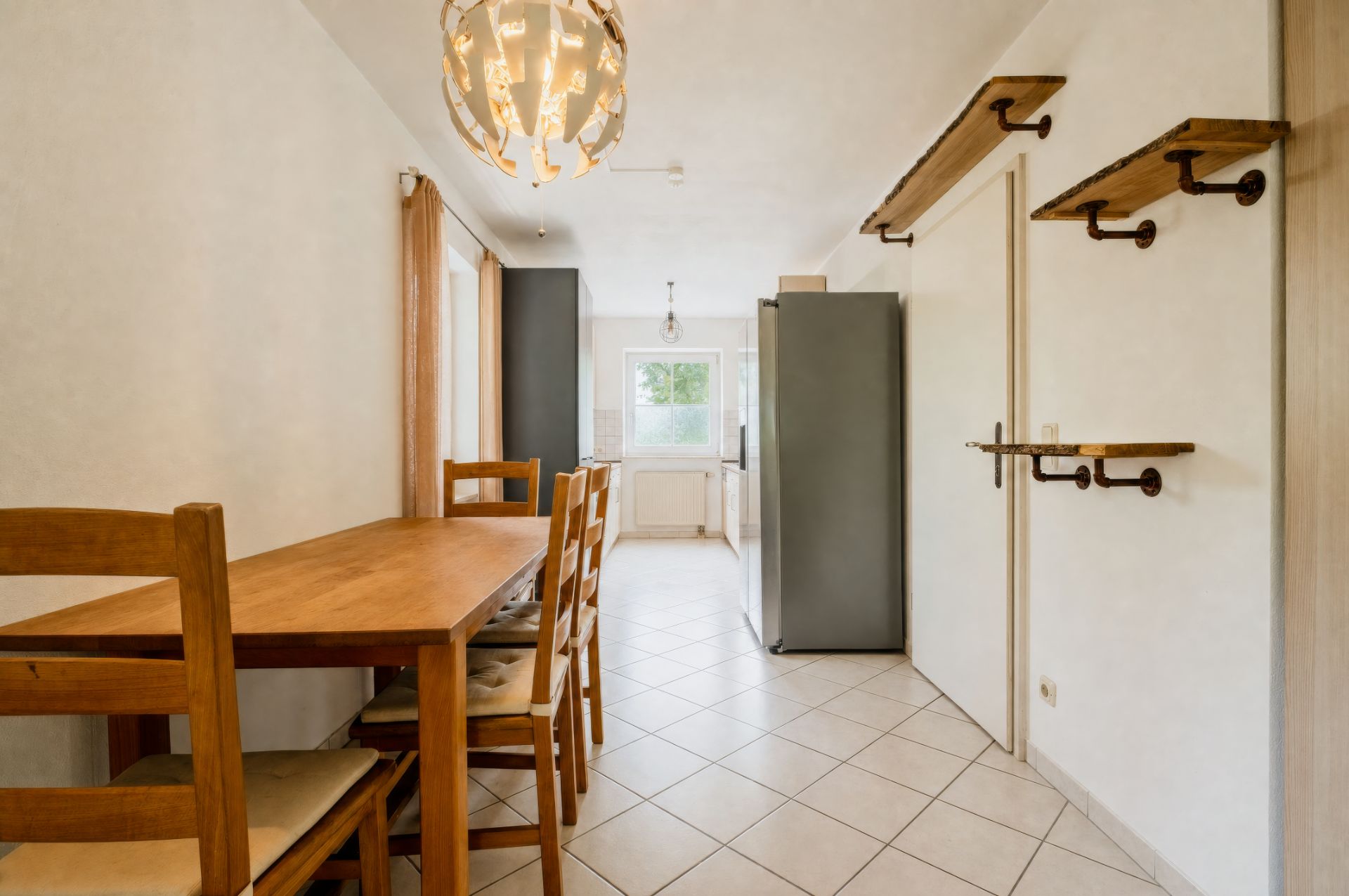 Helles Esszimmer mit Holztisch, vier Stühlen, Kühlschrank und Wandregalen, Blick auf Fenster.