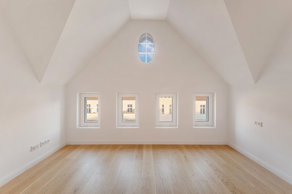 Helles Zimmer mit schräger Decke, Holzboden und fünf Fenstern an einer Wand.