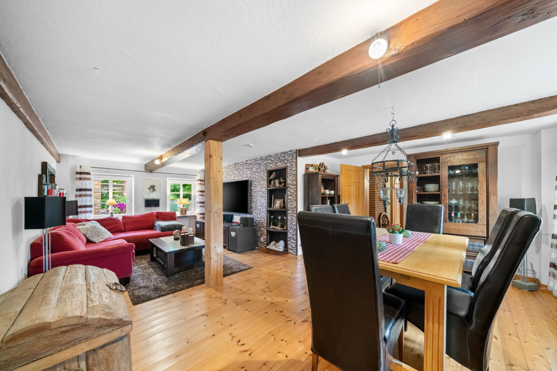 Helles Wohnzimmer mit Holzboden, rotem Sofa, Esstisch und sichtbaren Holzbalken an der Decke.