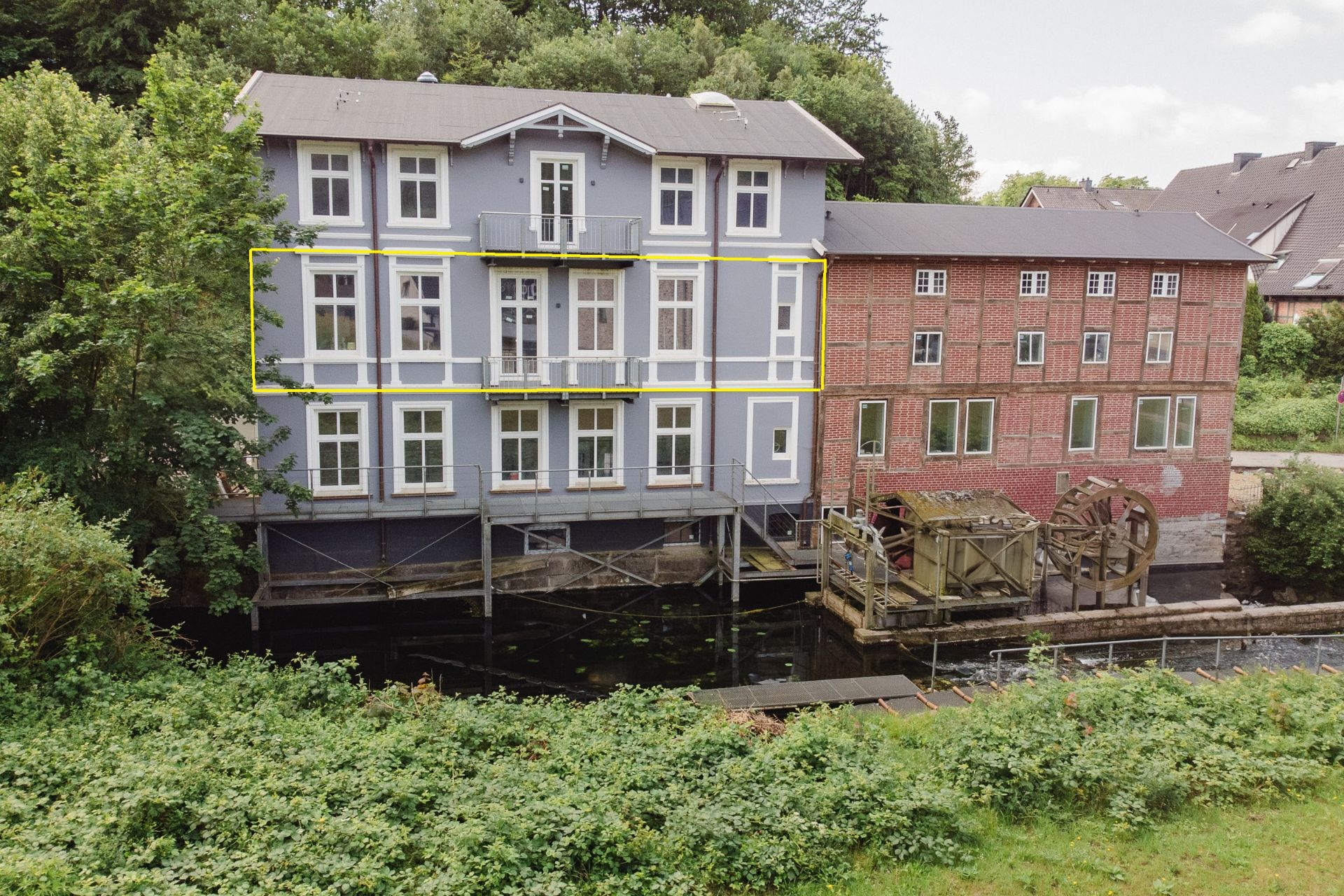 Zweistöckiges blaues Wohnhaus neben rotem Backsteingebäude an einem kleinen Fluss, mit Wasserrad.