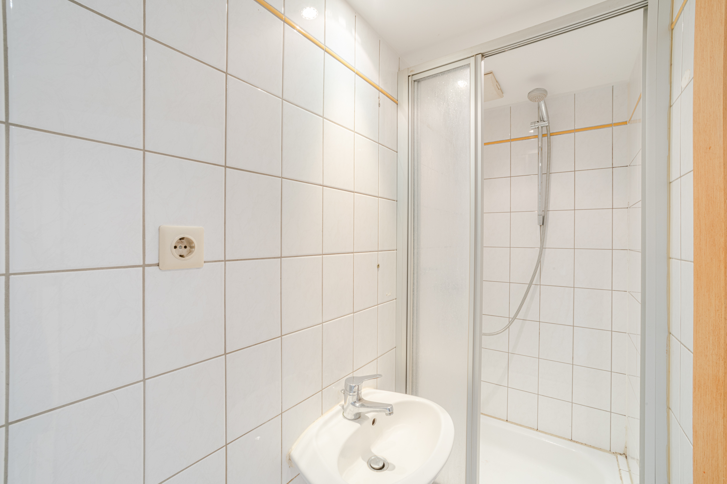 Kleines Badezimmer mit weiß gefliester Dusche, Waschbecken und Wandsteckdose.
