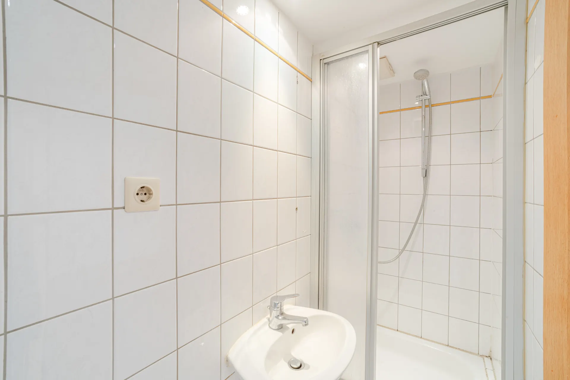 Kleines Badezimmer mit weiß gefliester Dusche, Waschbecken und Wandsteckdose.