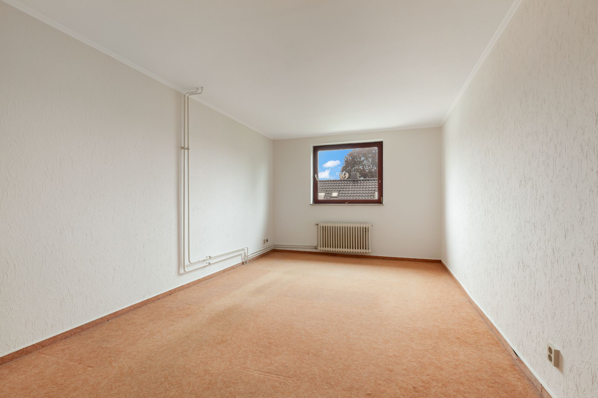 Leerer Raum mit braunem Teppich, weiß gestrichenen Wänden und einem Fenster mit Heizkörper darunter.