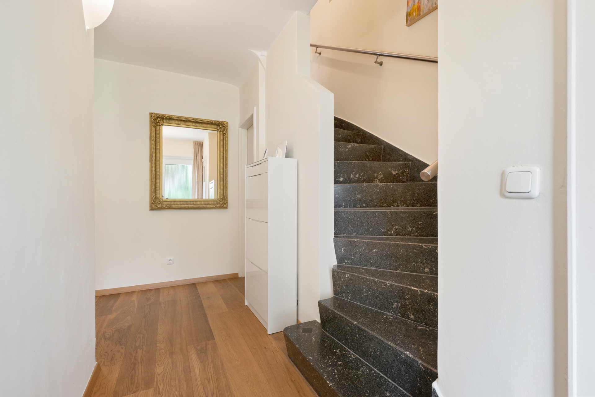Flur mit Holzfußboden, schwarzer Treppe und Wandspiegel in goldener Rahmen.