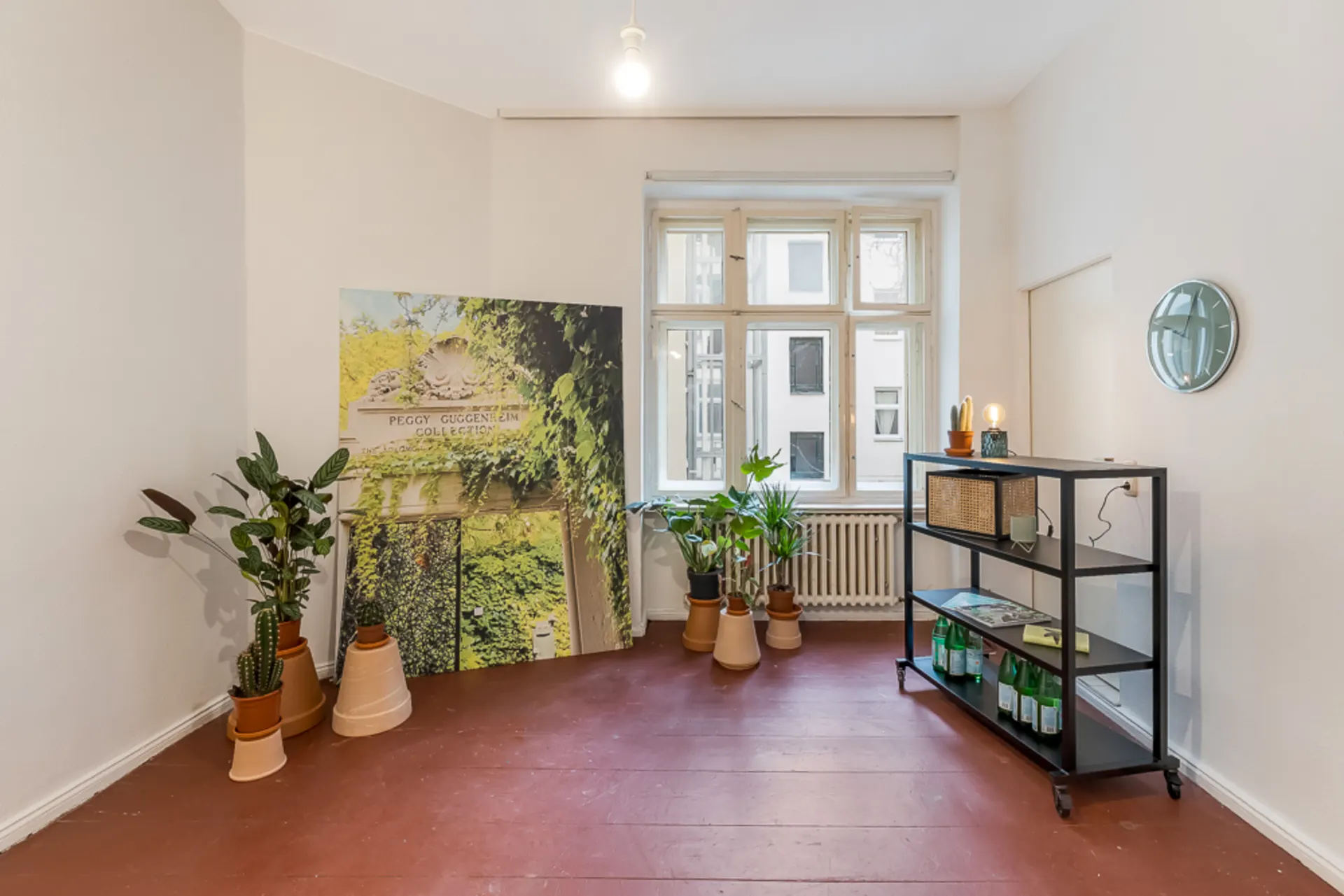 Heller Raum mit Pflanzen, einem Regal und großem Fenster, roter Boden.