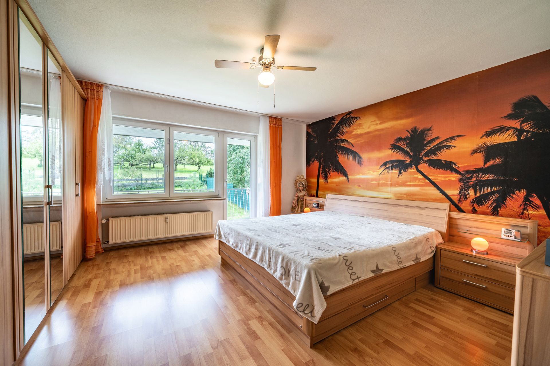 Helles Schlafzimmer mit großem Bett, Kleiderschrank und Wandbild mit Palmen bei Sonnenuntergang.